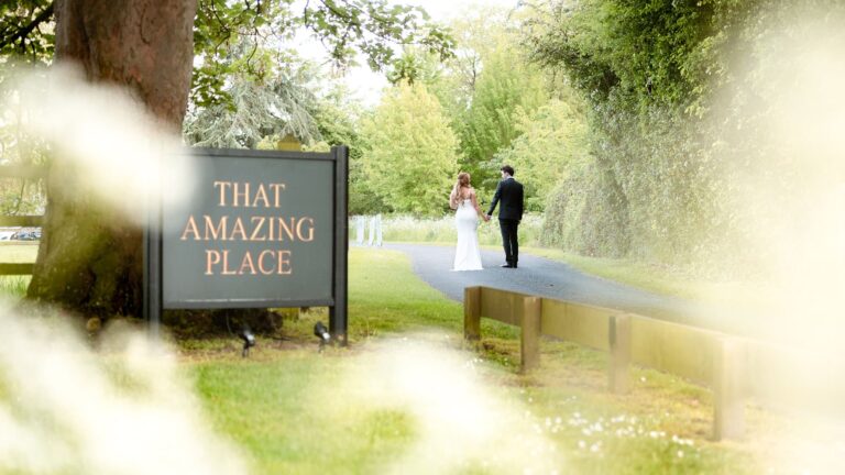 The tree-lined driveway with the venue sign, a couple walking in the distance, That Amazing Place