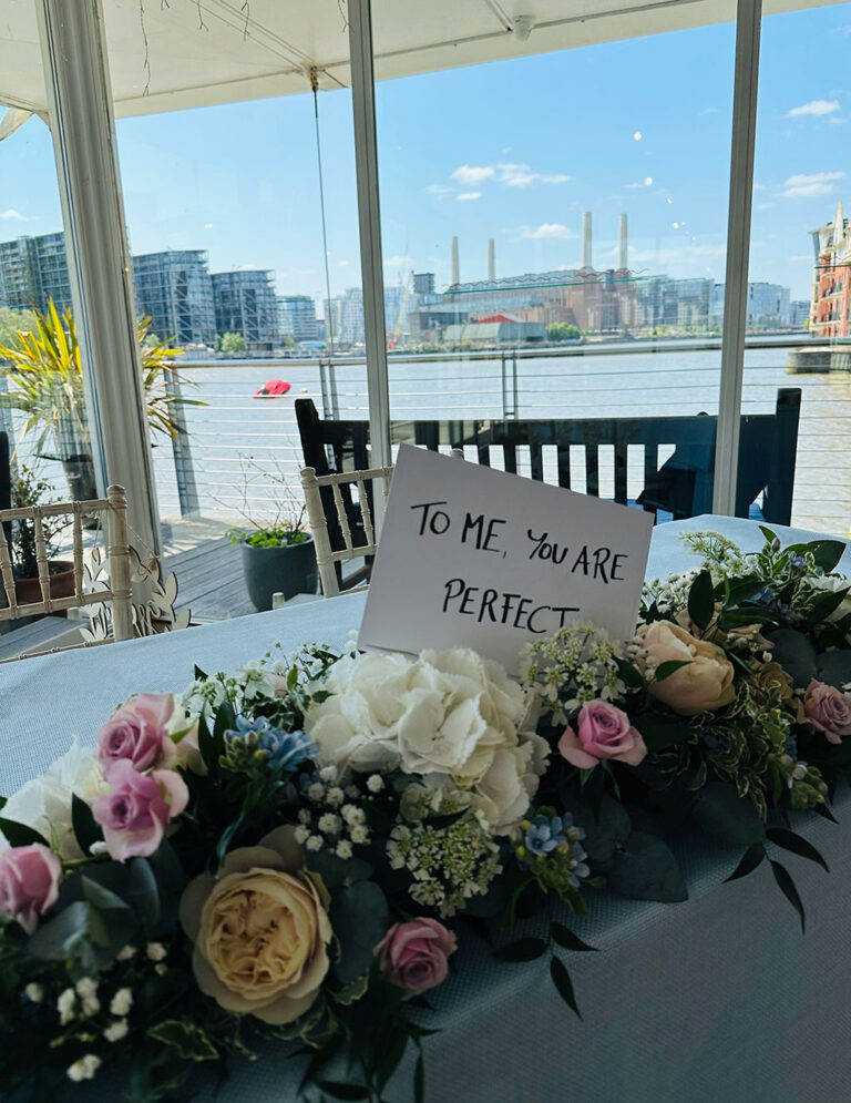 Head table flowers beside a river view window with a handwritten sign, The Admirable Crichton