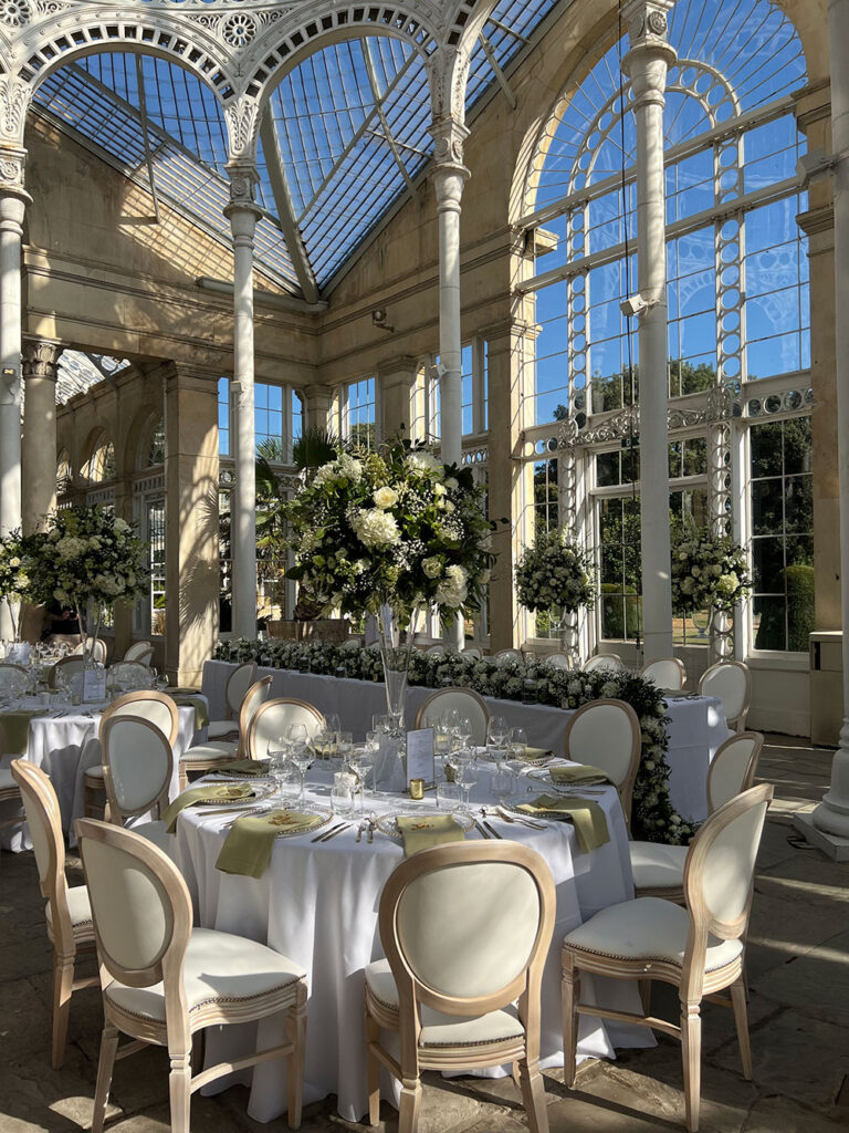 Round tables dressed in white with tall floral centrepieces inside a glasshouse, The Admirable Crichton