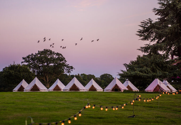 Bell tents on a lawn with festoon lighting at dusk, The Admirable Crichton