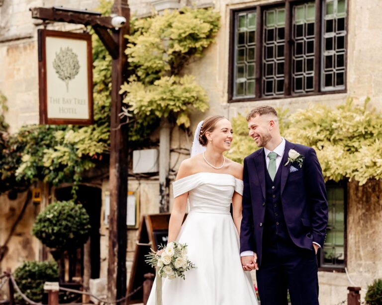 The couple outside the honey-stone entrance beneath the hotel's hanging sign and climbing greenery, The Bay Tree Hotel