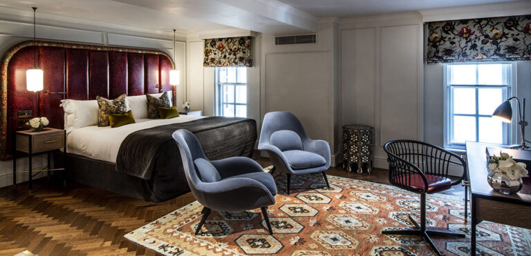 A suite bedroom with a curved red headboard, patterned rug and two armchairs by the window, The Bloomsbury