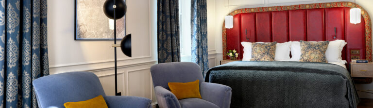 The bed and armchairs of a Luxury Studio Suite, with a red upholstered headboard and blue curtains, The Bloomsbury