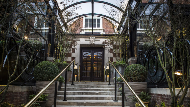 Stone steps and brass handrails lead up to the hotel's double front doors under a curved iron and glass canopy, flanked by clipped box and lanterns, The Bloomsbury
