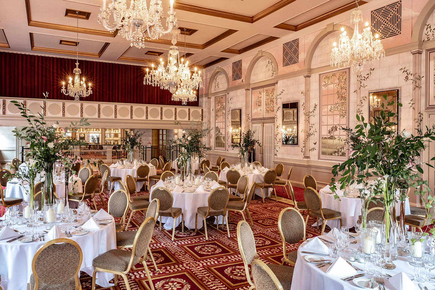 Queen Mary Hall laid with round tables for a wedding breakfast, under crystal chandeliers, with the new pale garden mural on the walls and the bar area beyond, The Bloomsbury