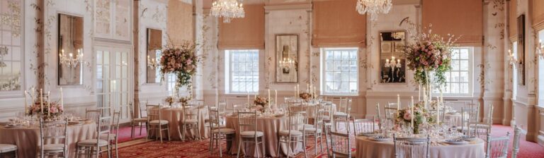 Queen Mary Hall dressed in blush tones, with tall floral centrepieces, candles and chiavari chairs around round tables, The Bloomsbury