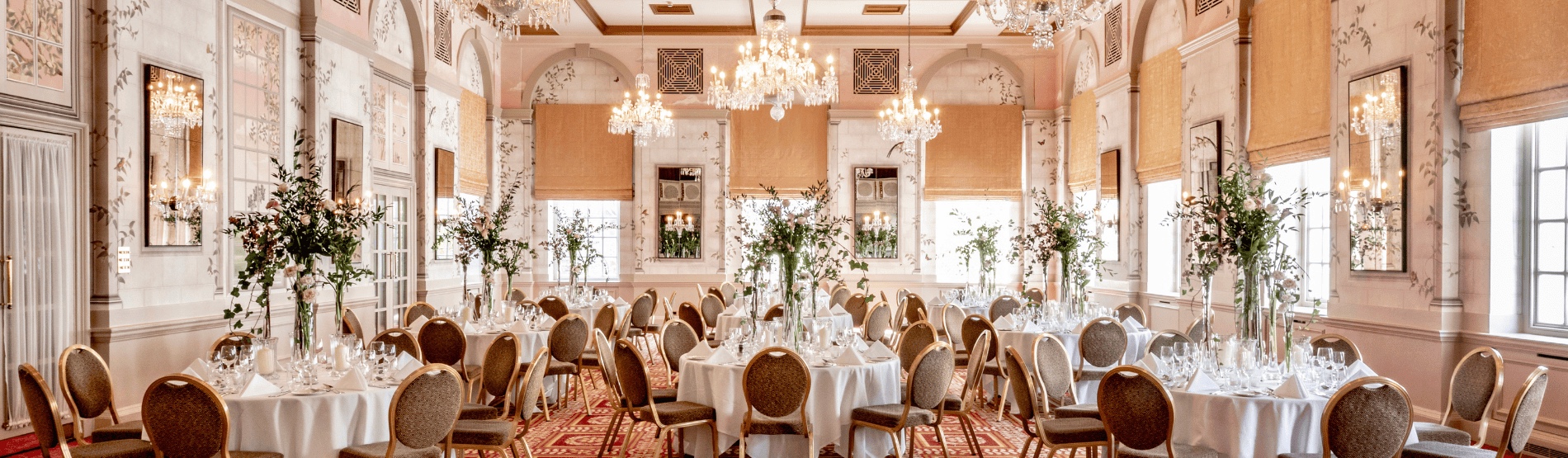 A wide view of Queen Mary Hall set for dinner, with tall window blinds, mirrored panels and greenery on every table, The Bloomsbury
