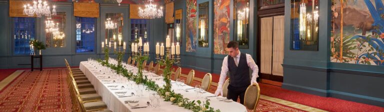 A long banqueting table in George V Hall with candelabra and a trailing greenery runner, as a member of staff sets the chairs, The Bloomsbury