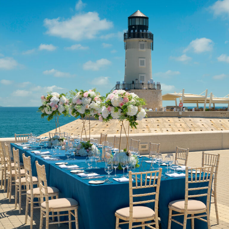 A long dining table set on the breakwater with the lighthouse behind, The Chedi Luštica Bay