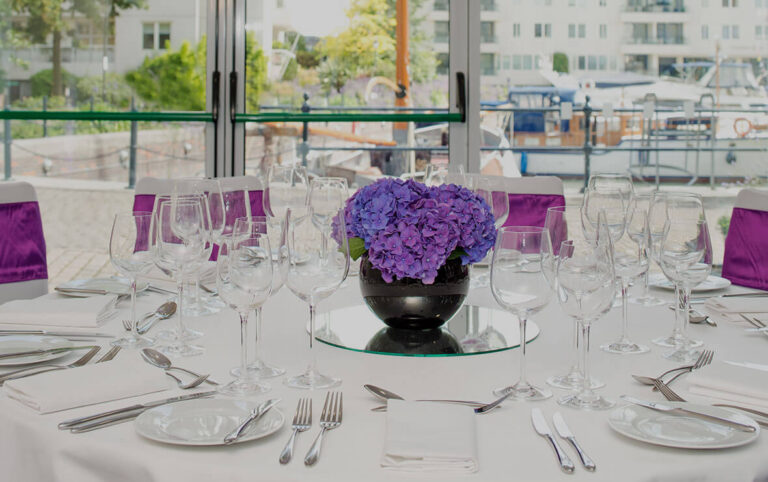 A laid round table with hydrangeas and glassware in front of glass doors onto the marina, The Chelsea Harbour Hotel