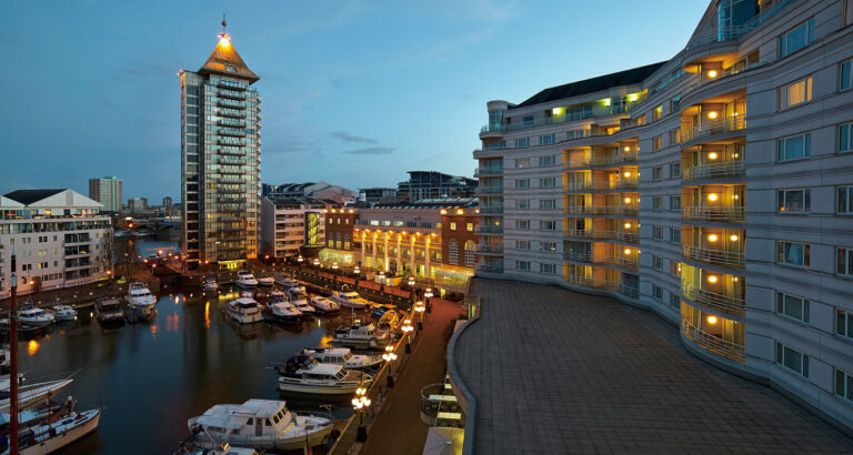 The curved hotel facade at dusk, lit windows running along the marina, with moored boats and the Belvedere tower behind, The Chelsea Harbour Hotel