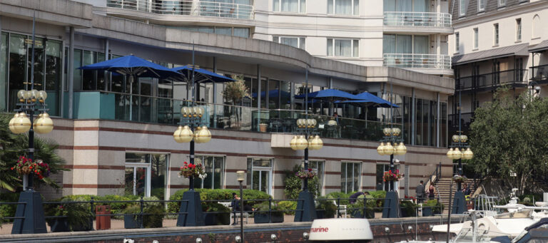 The hotel frontage on the harbour walk, with the restaurant terrace under blue parasols and lanterns along the quay, The Chelsea Harbour Hotel