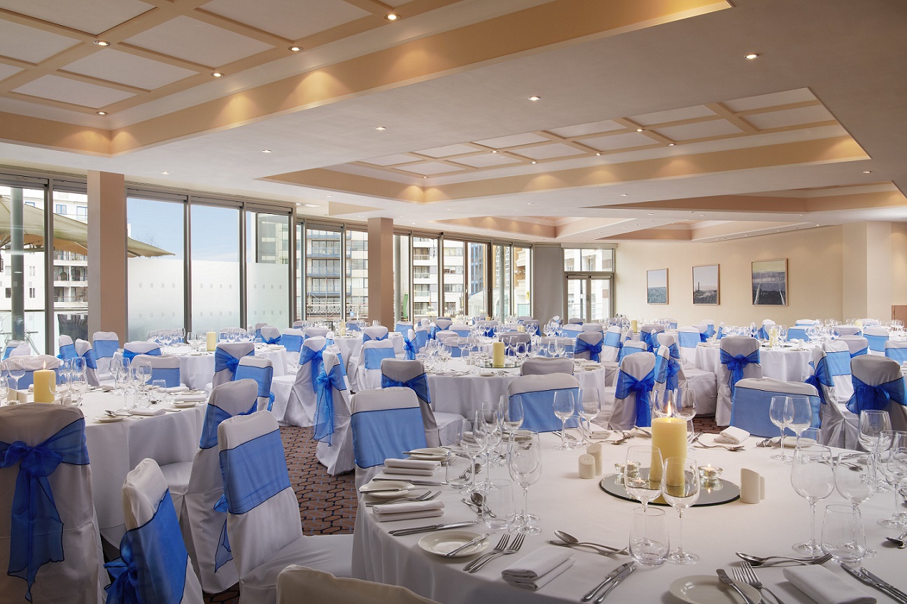 The Drakes Suite on the lobby level laid for a wedding dinner, round tables with blue sashes and floor-to-ceiling windows, The Chelsea Harbour Hotel