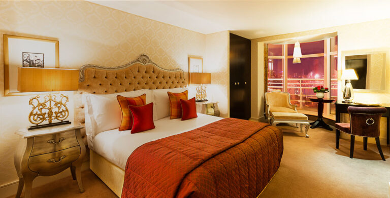 A penthouse bedroom with a buttoned headboard, orange throw and a seating area by the window, The Chelsea Harbour Hotel