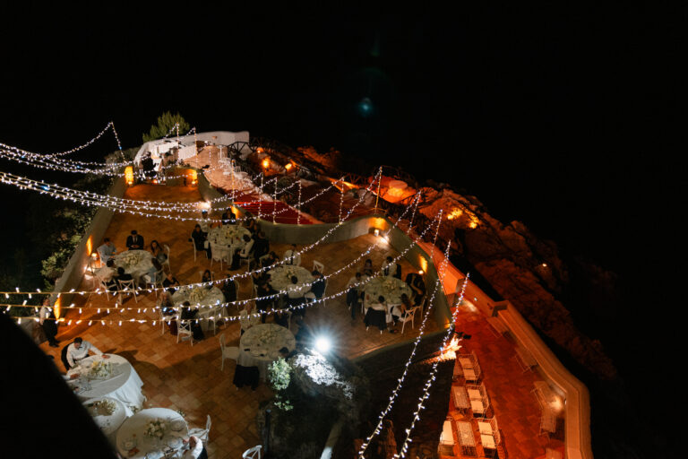 Looking down on a cliffside terrace reception at night, lit with festoon lights, The Dream Wedding Lab