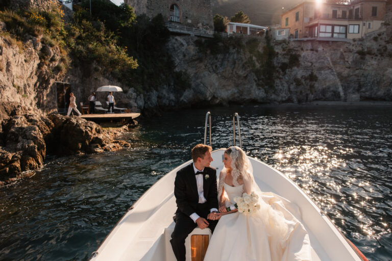 The couple seated at the bow of a boat beneath rocky cliffs, sunlight on the water, The Dream Wedding Lab