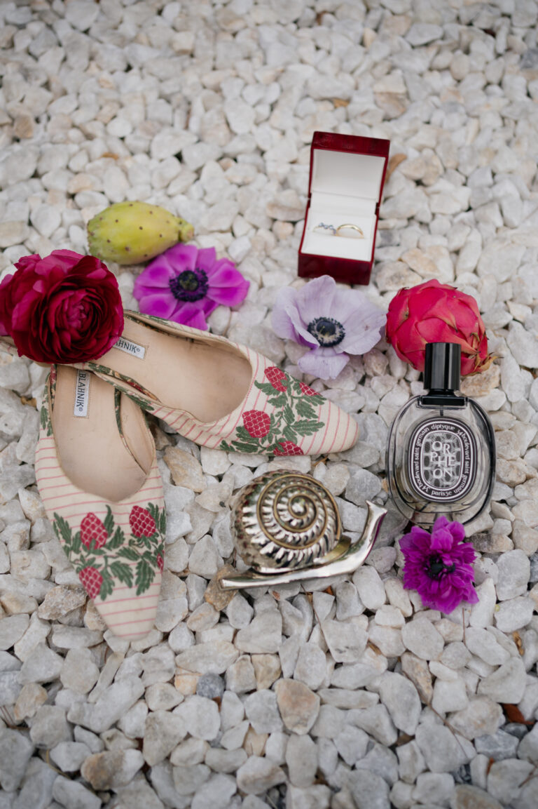 A ring box, embroidered shoes, perfume and flowers laid out on white pebbles, The Dream Wedding Lab
