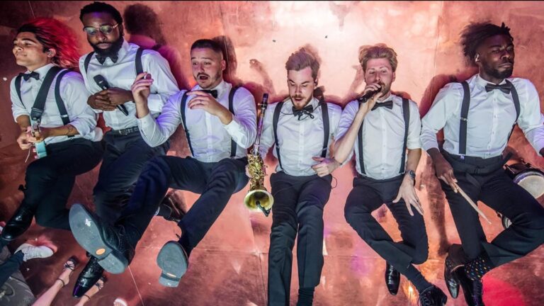 Six band members mid-jump in white shirts and braces against a coloured backdrop, The Function Band