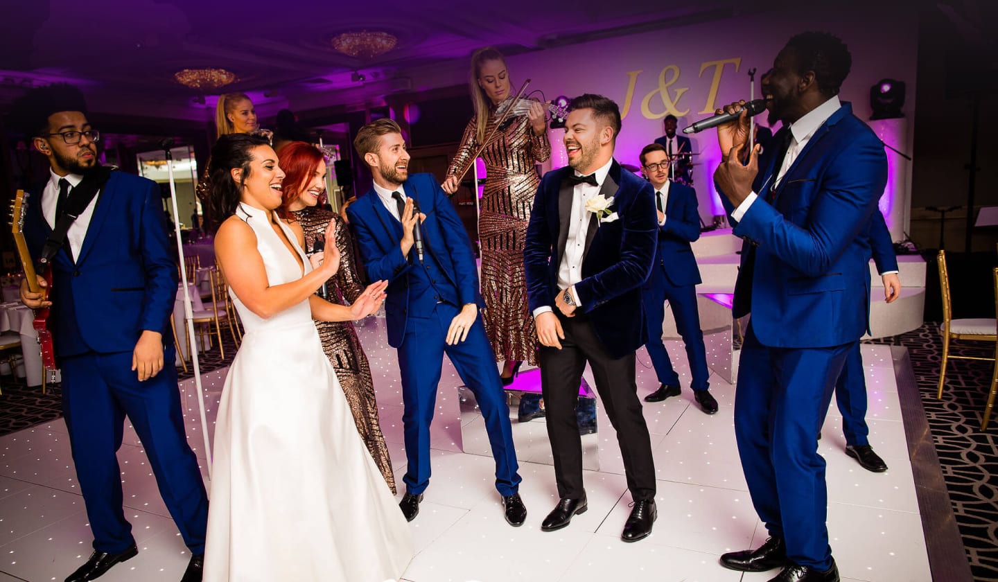 Vocalists and the band with the bride and groom on a lit dance floor at a hotel wedding, The Function Band
