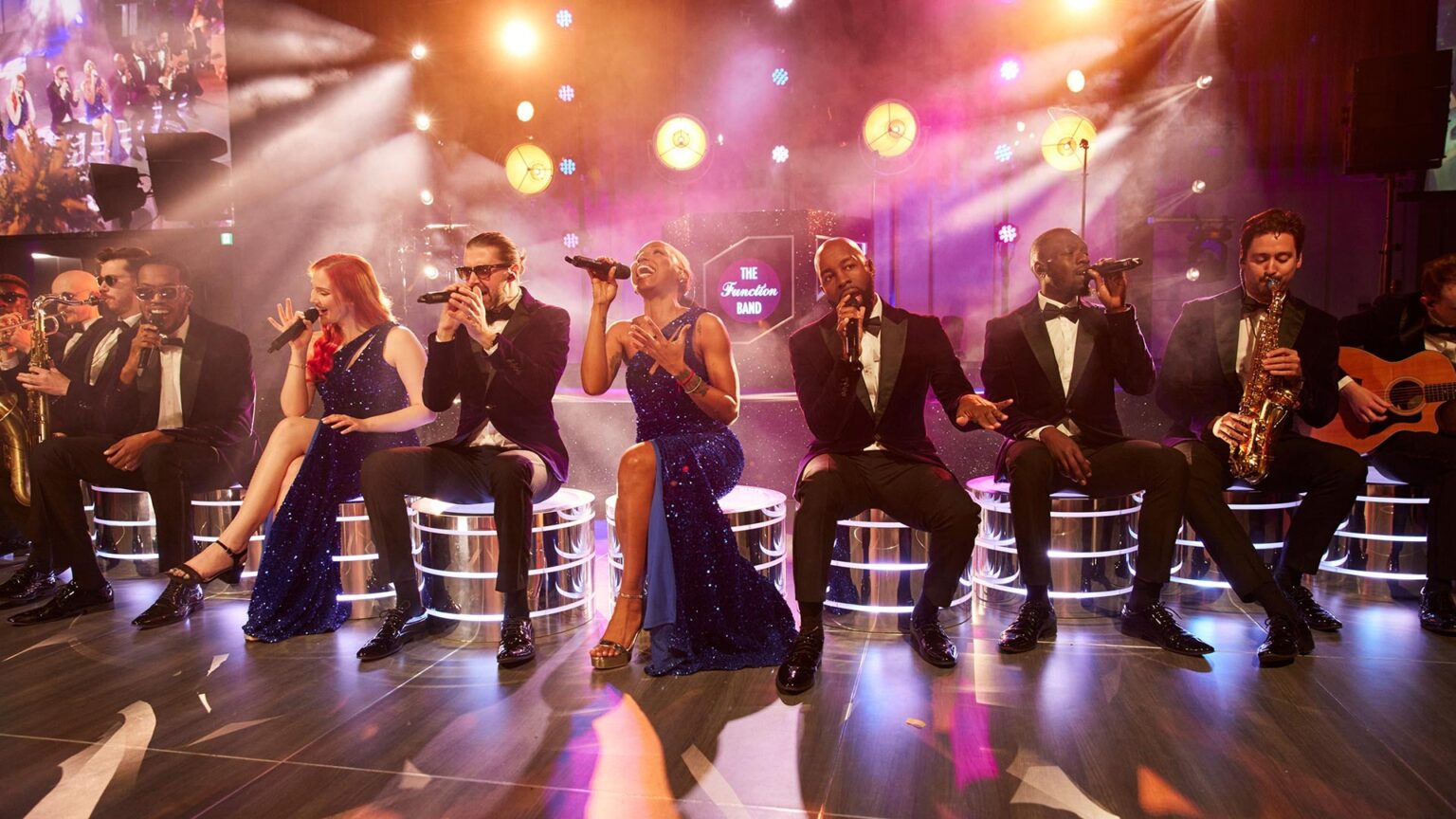 The full band and vocalists on lit stools with a haze-filled stage show