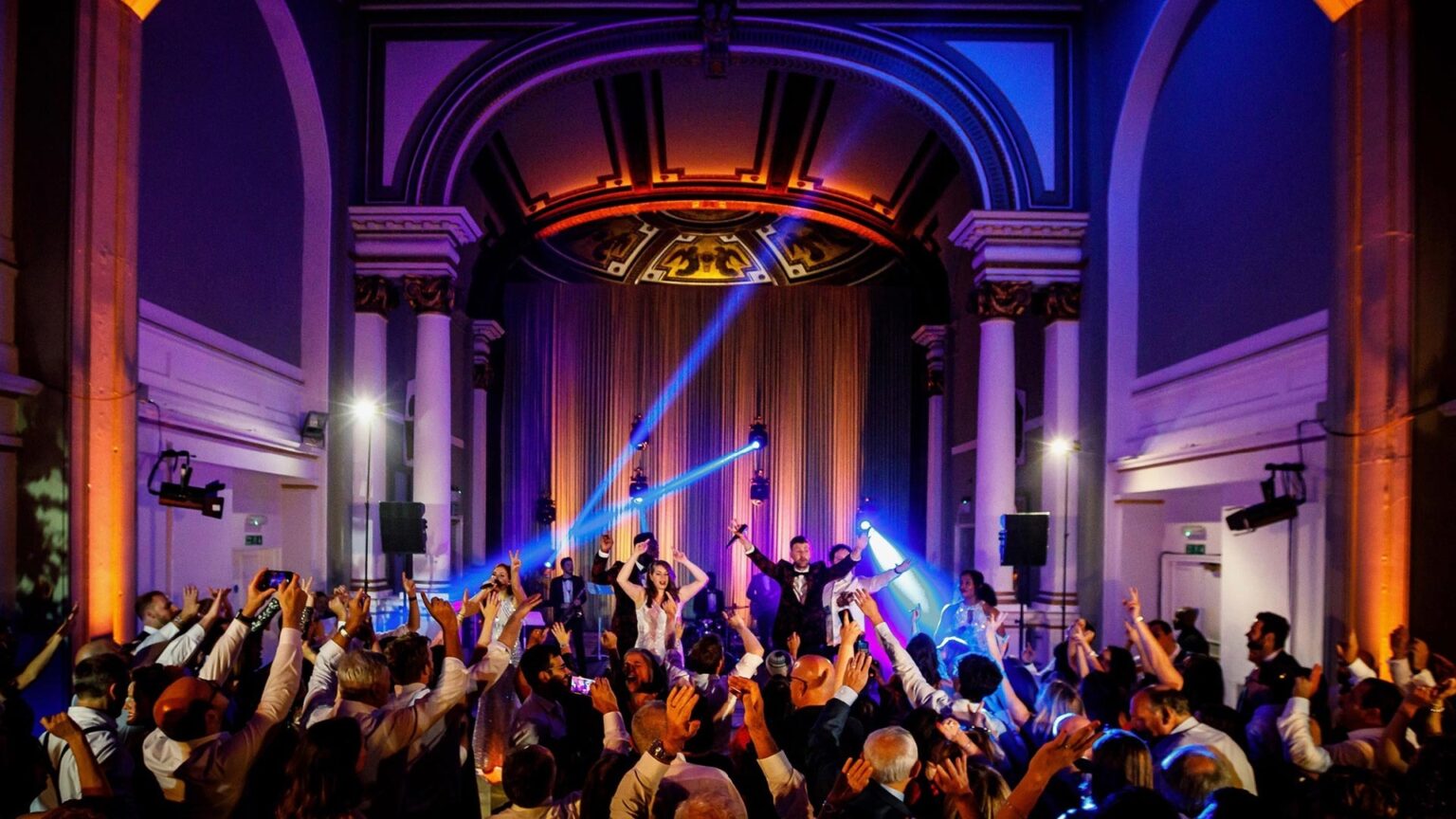 A crowd with raised arms in a grand domed hall lit with blue and amber beams
