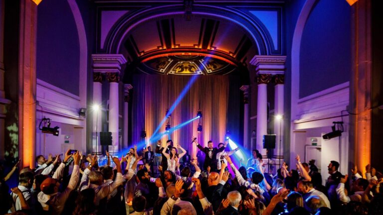 Crowd with raised arms in a grand domed hall lit with blue and amber beams, The Function Band