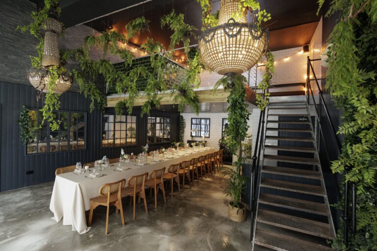 The glasshouse dining room, greenery and pendant lights above a long laid table, The Greenhouses