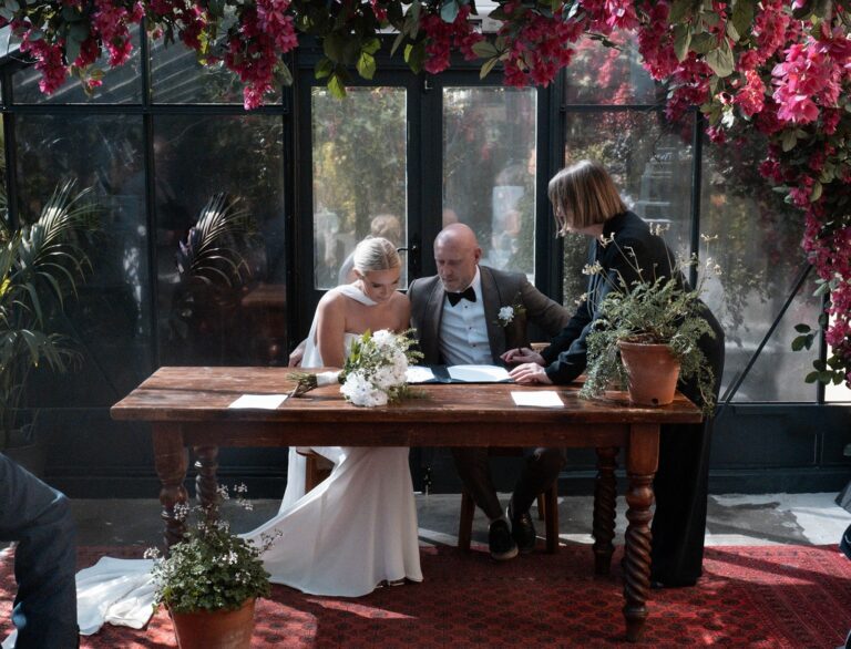 The couple signing the register at a wooden table beneath bougainvillea, The Greenhouses