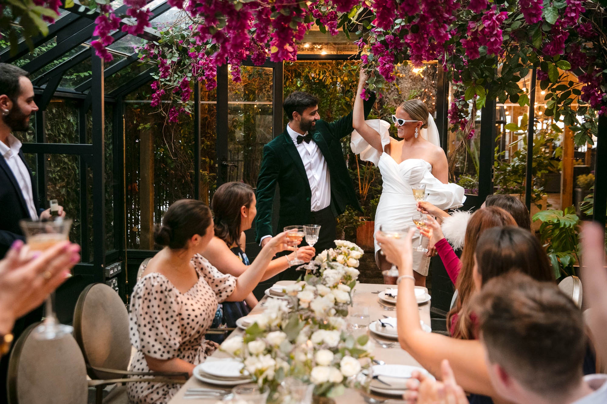 Guests applauding as the couple stand at the table, greenery overhead, The Greenhouses