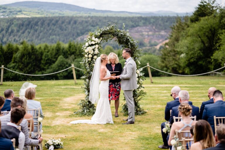 A couple exchanging vows under a floral arch on the lawn, with the wooded valley as the backdrop, The Horn Of Plenty