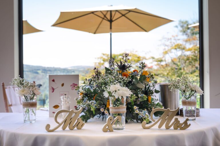 The couple's table with wild flowers and Mr and Mrs letters, under parasols on the terrace beyond the glass, The Horn Of Plenty