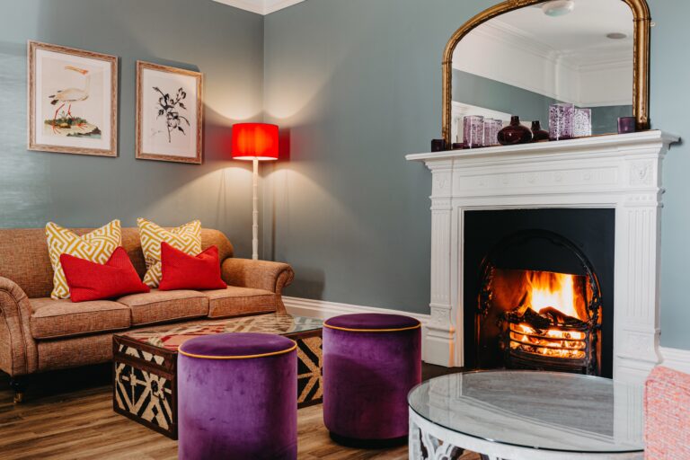 The drawing room with a lit log fire, velvet stools and a gilt mirror over the mantelpiece, The Horn Of Plenty
