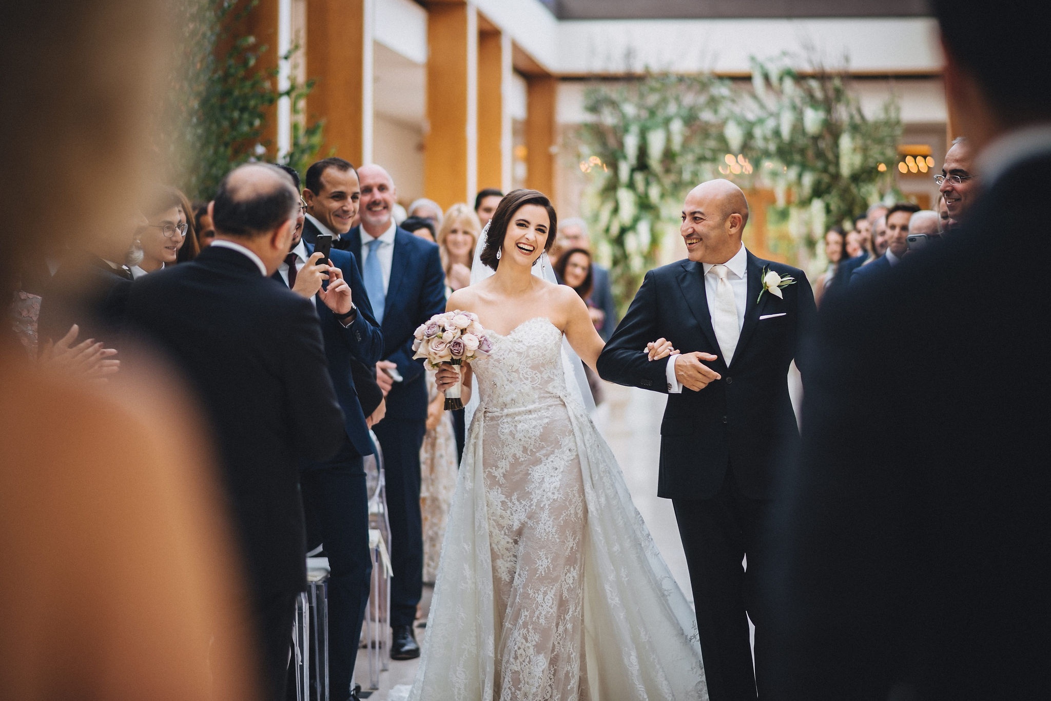 A bride walking down the aisle in Palm Court, guests seated beneath the glass roof, The Hurlingham Club