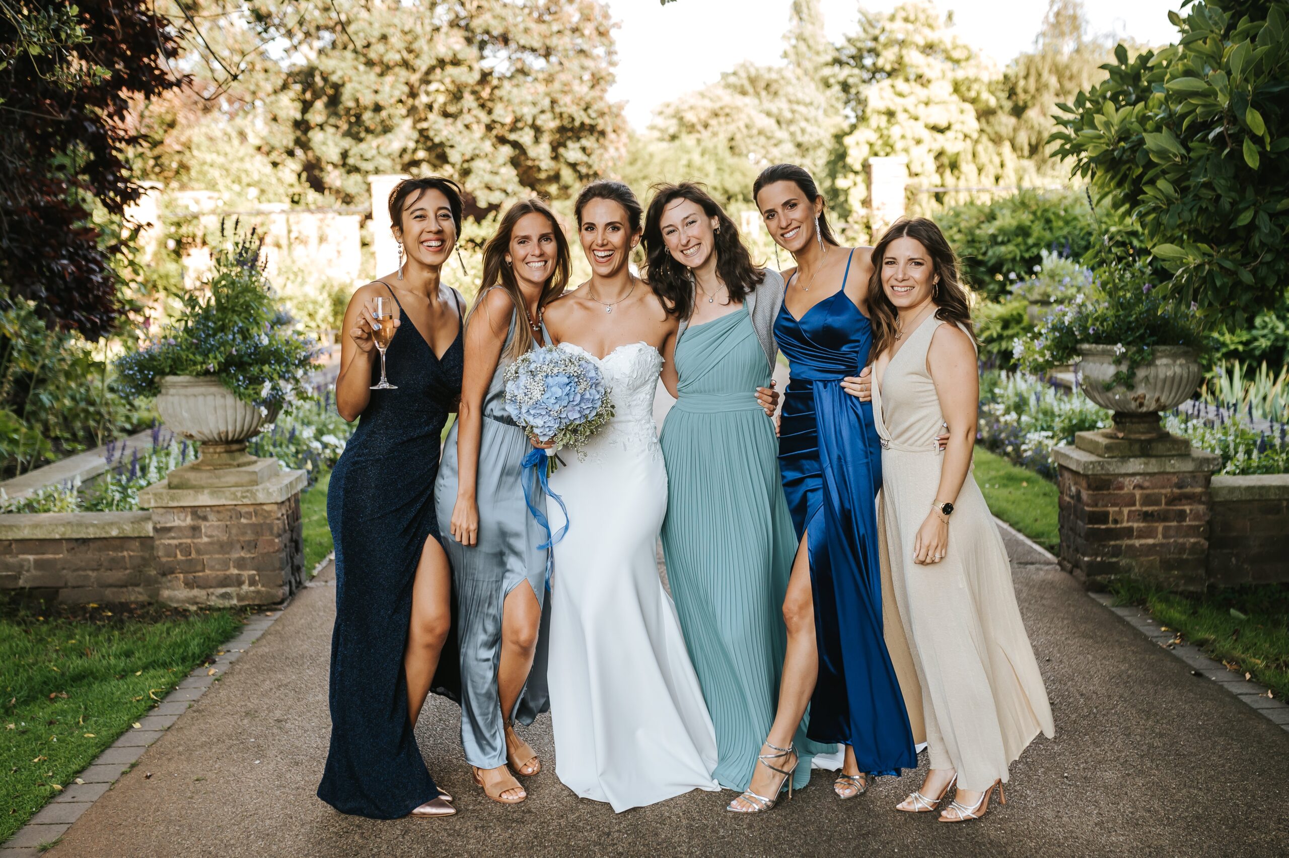 A bride and her bridesmaids in blue and green on the path through the Sunken Garden, The Hurlingham Club