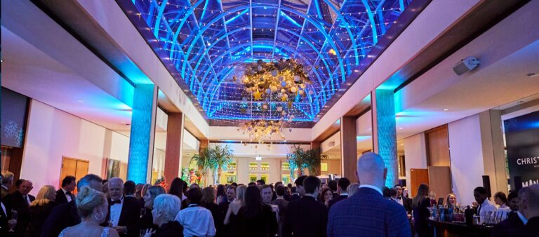 A drinks reception filling Palm Court under the lit glass dome and chandelier, The Hurlingham Club