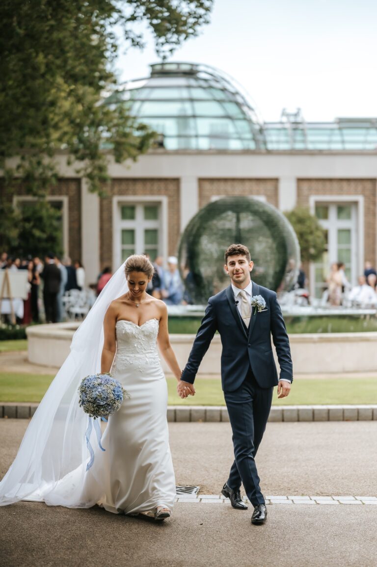 A bride and groom walking hand in hand past the glass dome of Palm Court, The Hurlingham Club