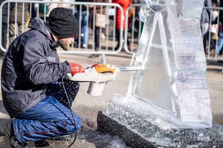 A sculptor shaping a tall block of ice beside the barriers at a live carving, The Ice Box