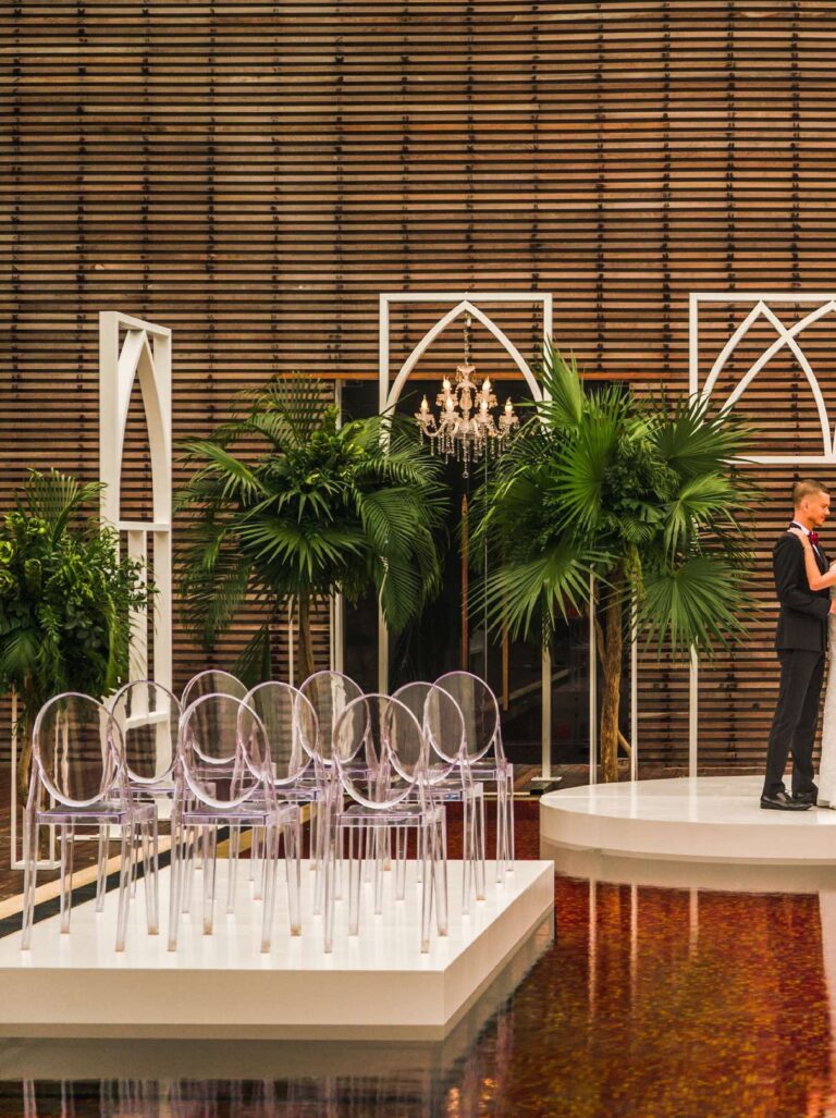 Ceremony stage with clear chairs, arched flower frames and palms, The Library