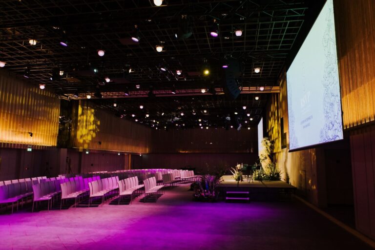 The Ballroom set with rows of chairs facing a flower-dressed stage and projection screen, The Londoner