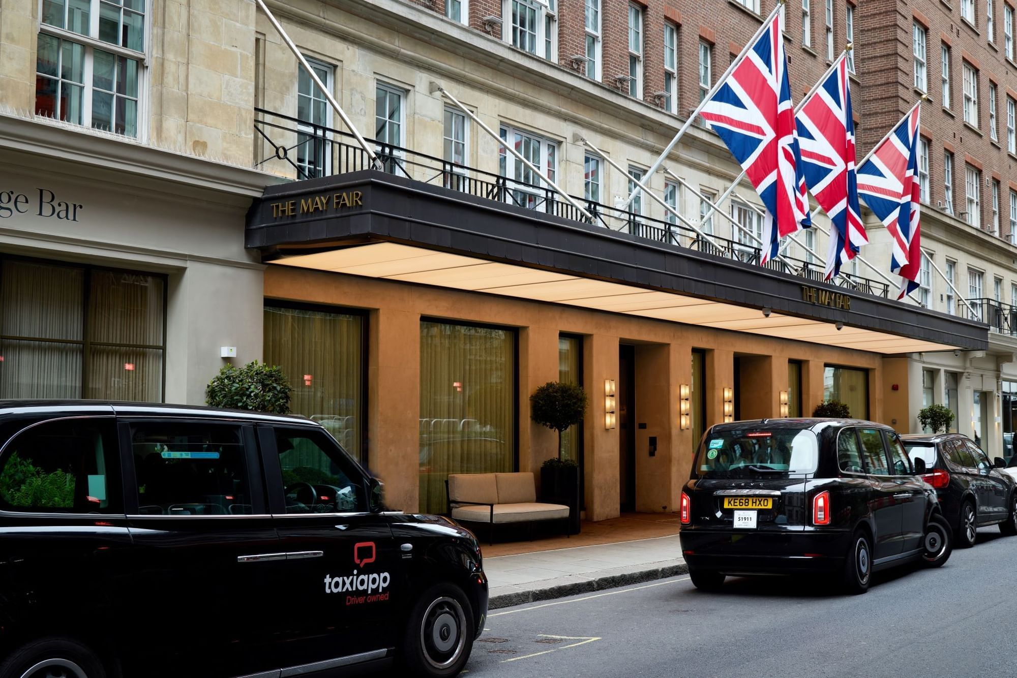The canopied main entrance on Stratton Street by day, with flags above and black cabs at the kerb, The May Fair