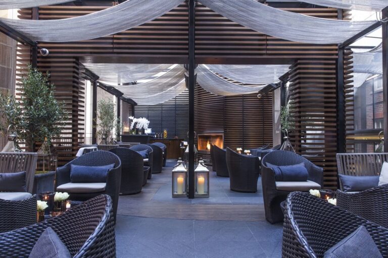 The May Fair Terrace by day, a sheltered courtyard with wicker seating, lanterns and a fireplace under fabric canopies, The May Fair