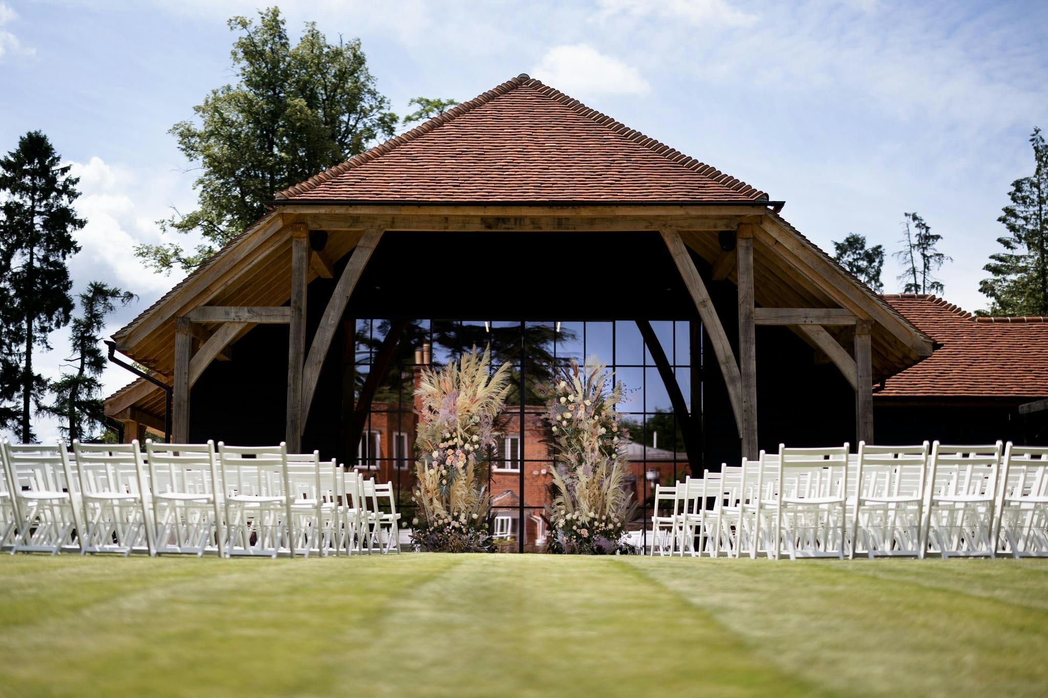 White chairs set on the lawn for an outdoor ceremony, facing the barn's glazed gable and a pampas-grass floral arch, The Post Barn