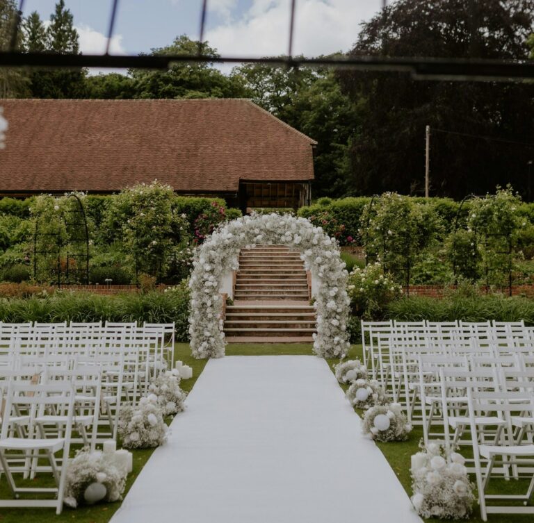 A white aisle leading to a floral arch at the foot of brick garden steps, with the barn roof behind, The Post Barn