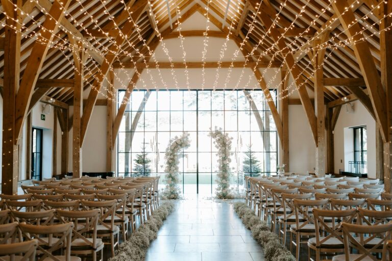 The barn set for an indoor ceremony under oak trusses hung with fairy lights, with floral arches at the glazed end, The Post Barn