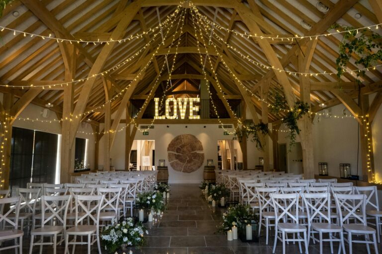 Rows of white chairs and a candlelit aisle in the barn, with illuminated LOVE letters on the mezzanine, The Post Barn