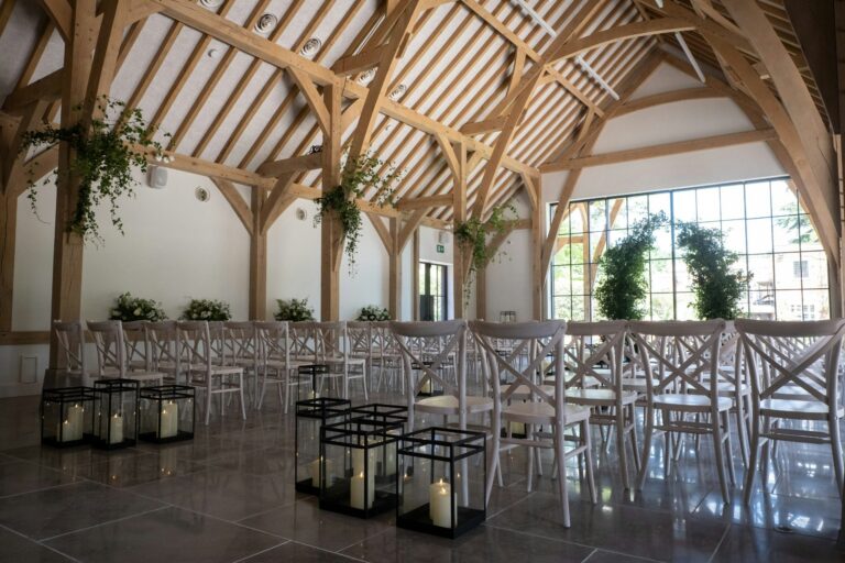 The barn set for a ceremony with glass lanterns along the aisle and greenery in the oak frame, The Post Barn