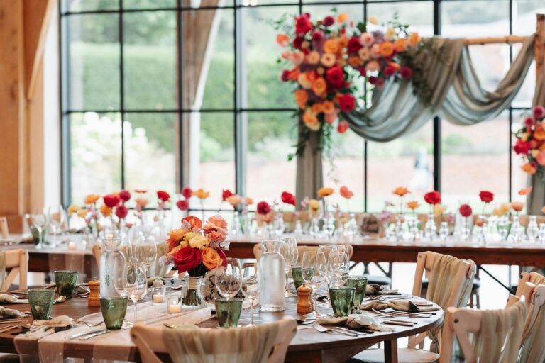 A round table laid with green glassware and orange and pink flowers, a floral arch on the window behind, The Post Barn