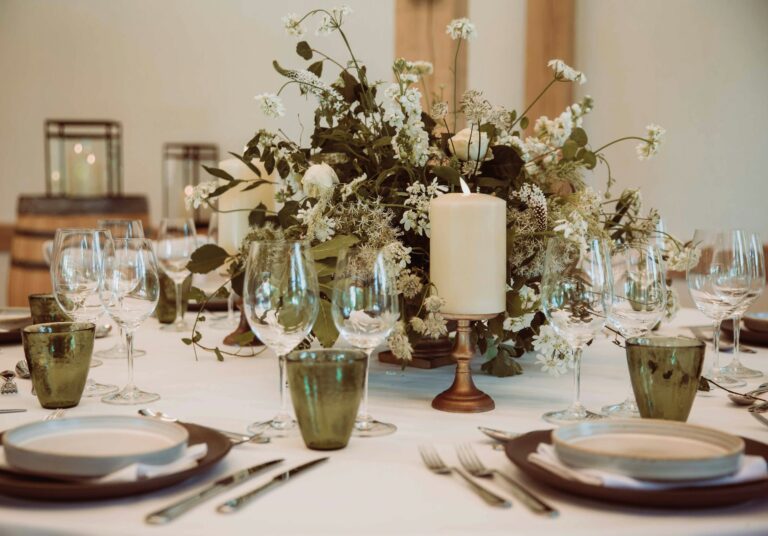 A table setting with a wild-flower centrepiece, pillar candle and green-tinted glasses, The Post Barn