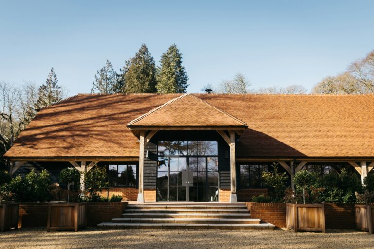 The front of the barn on a clear day, with its oak porch, glazed doors, stone steps and timber planters below a clay-tiled roof, The Post Barn