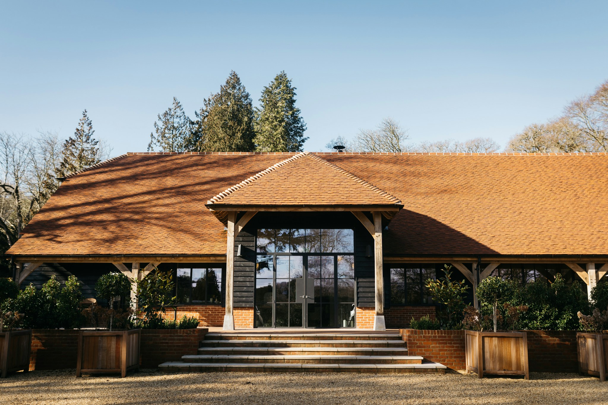The front of the barn on a clear day, with its oak porch, glazed doors, stone steps and timber planters below a clay-tiled roof, The Post Barn